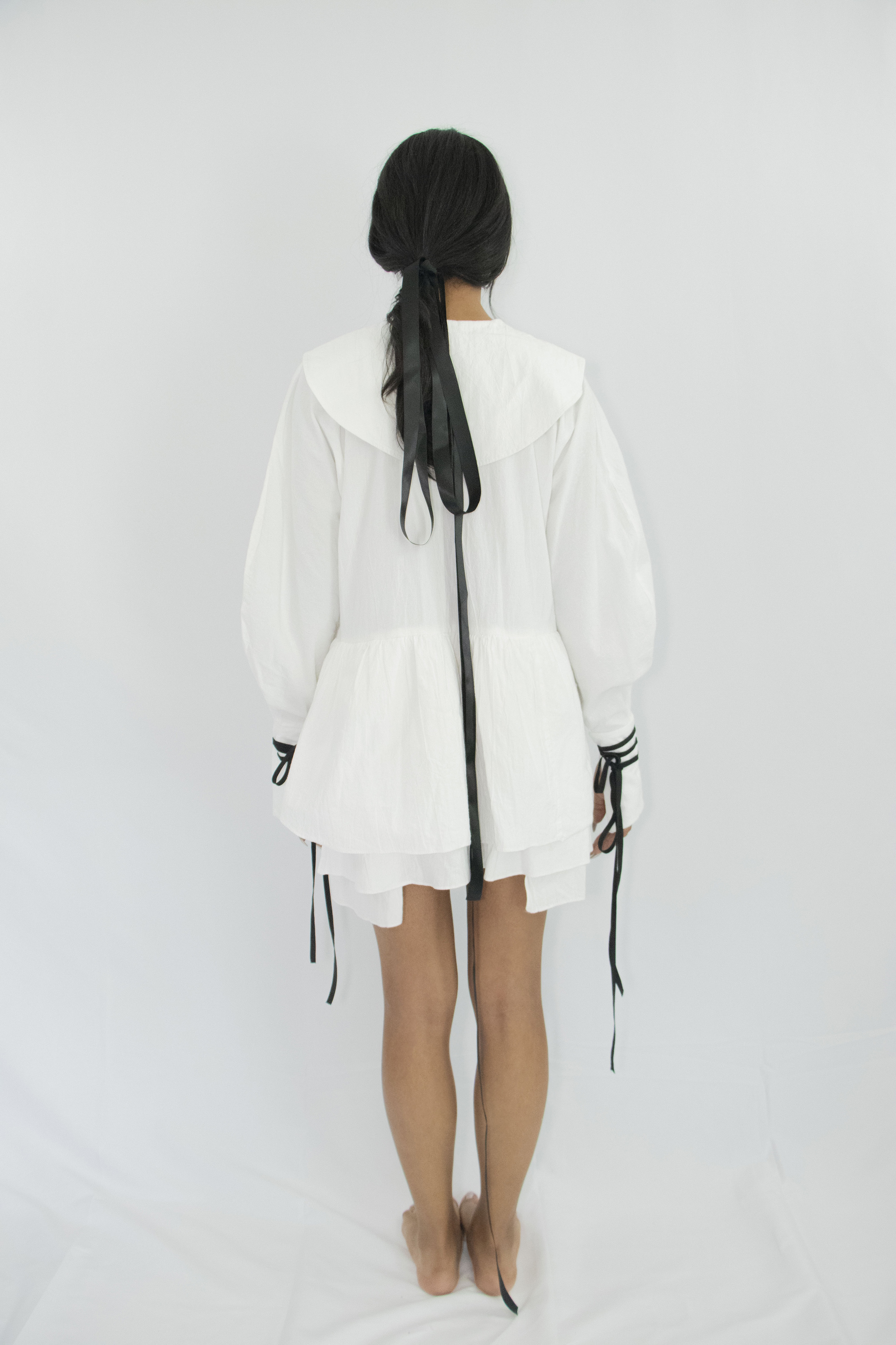 Woman wearing white organic cotton short dress with kimono sleeves, large cuffs with attachable contrasting black straps, pilgrim collar and gathered panels at the waist