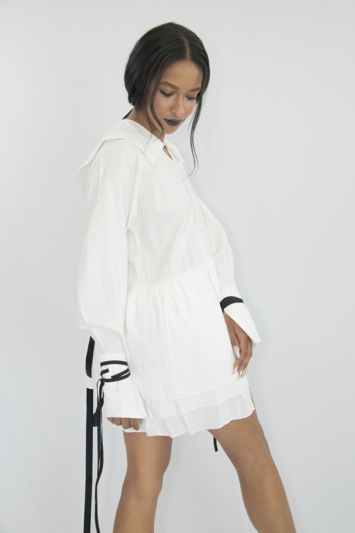Woman wearing white organic cotton short dress with kimono sleeves, large cuffs with attachable contrasting black straps, pilgrim collar and gathered panels at the waist