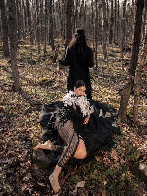 Woman laying on rock in the woods wearing a zero waste black and white fringed cape with man standing in the background